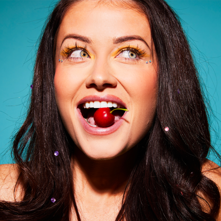 A young woman holding a cherry in her mouth against a teal backdrop.