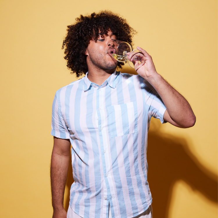 Curly afro haired man drinking white wine. The backdrop is a solid yellow and his shirt is white/light blue striped.