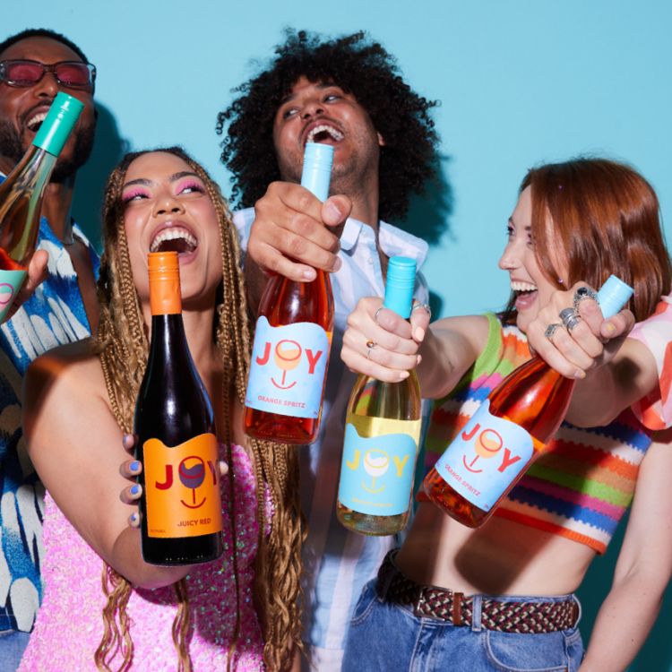 People in their twenties holding different bottles of JOY wine. The people are diverse and there's a solid blue backdrop.