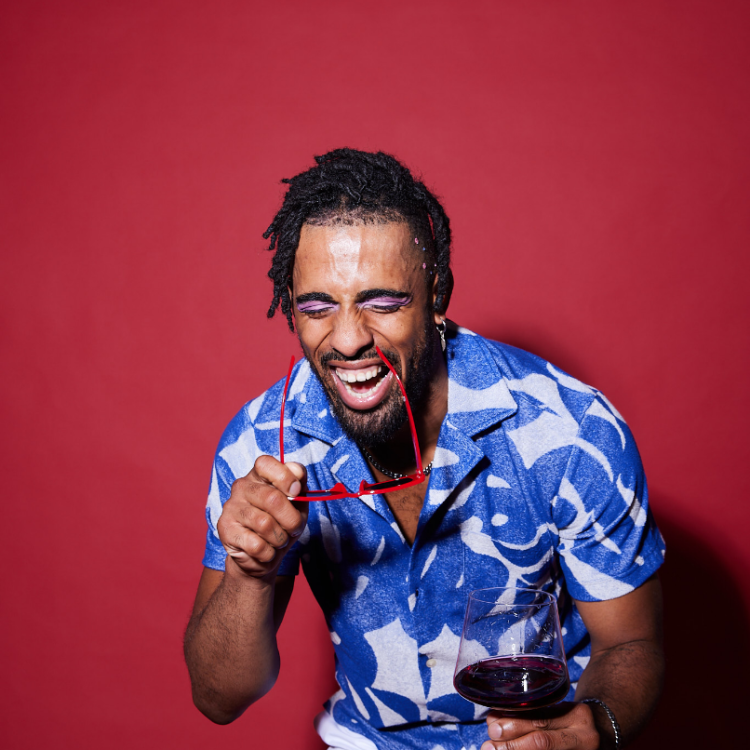 Man with afro hair and pink eyeliner in a blue patterned shirt laughing with his eyes closed while holding a glass of red wine.