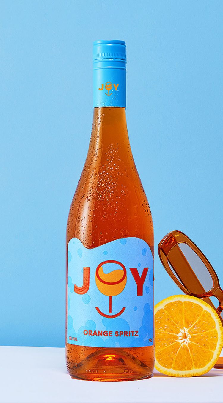 A bottle of JOY Orange Spritz against a blue backdrop. In the foreground is half an orange and a pair of sunglasses.
