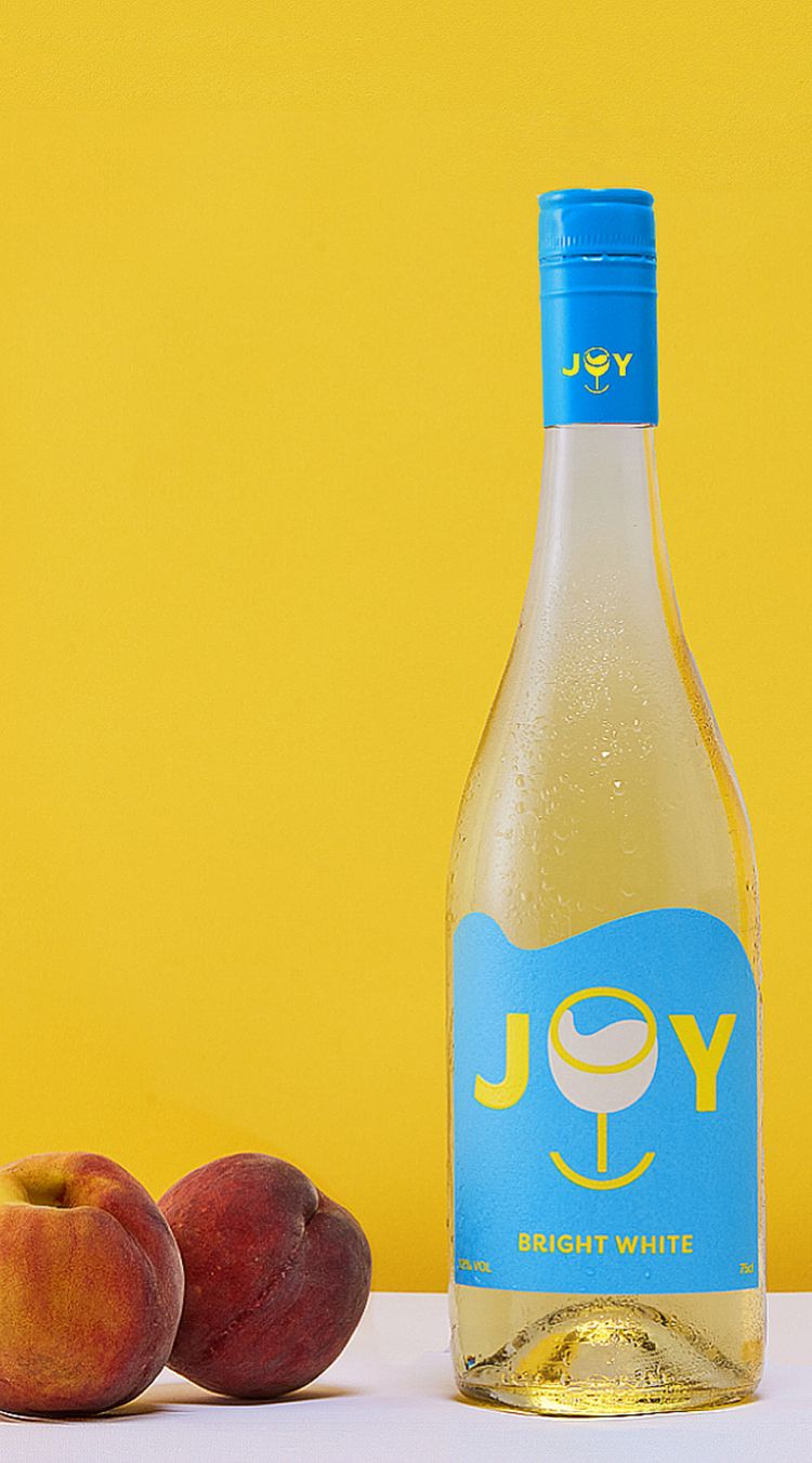 A bottle of JOY Bright White against a yellow backdrop. In the foreground are a couple of peaches.