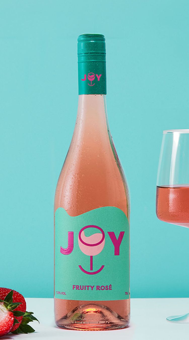 A bottle of JOY Fruity Rosé against a light blue backdrop. In the foreground are some strawberries.
