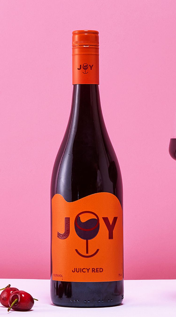 A bottle of JOY Juicy Red against a soft red, almost purple, backdrop. There's a glass poured next to it. In the front-left foreground are some cherries.